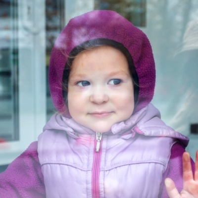 Criança com casaco lilás e capuz cor vinho atrás de janela de vidro