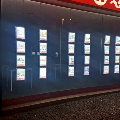 Vitrine iluminada de agência imobiliária com anúncios em acrílico transparente e faixa vermelha com texto branco