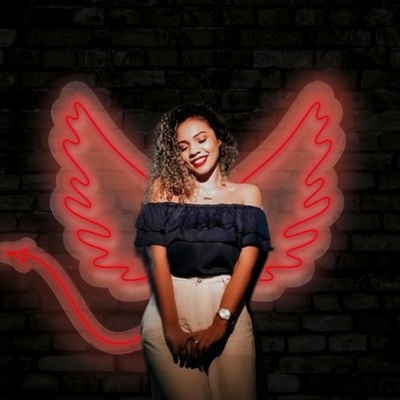 Jovem sorridente com blusa preta e calças brancas diante de parede de tijolos com asas vermelhas iluminadas atrás