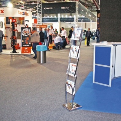 Estandes de feira com produtos, logos 'tecôfix' e 'OLIPAL', suporte metálico com folhetos, balcão branco com azul e banco alto em tapete azul