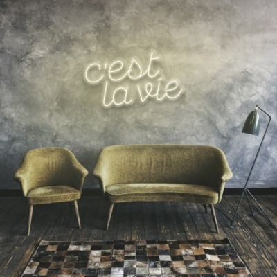 Sofá e poltrona verde-oliva com tapete geométrico e candeeiro junto a parede cinzenta com letreiro luminoso