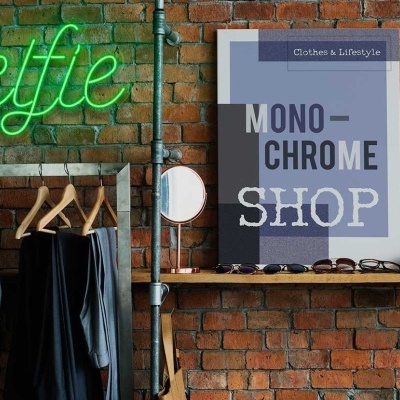 Parede de tijolos com placa néon verde 'selfie', cabides com roupa escura, e cartaz 'MONO-CHROME SHOP' com acessórios.