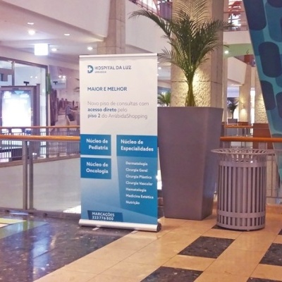 Banner informativo da HOSPITAL DA LUZ num centro comercial com vasos e contentor de lixo ao redor