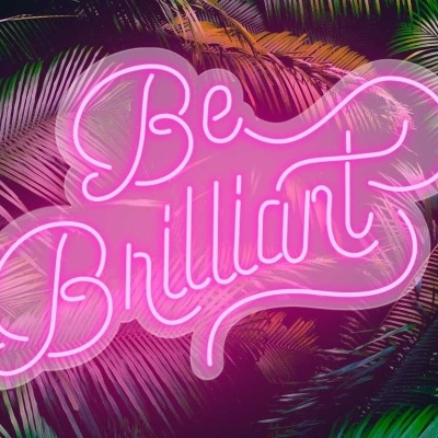 Placa de néon rosa com texto Be Brilliant sobre folhas tropicais verdes.