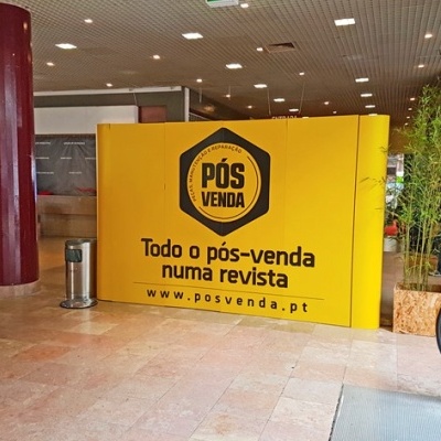 Painel publicitário amarelo com texto preto num espaço interior com colunas vermelhas.