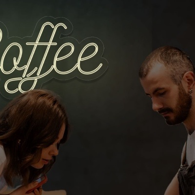Neon amarelo com a palavra 'Coffee' contra parede escura, duas pessoas olhando para baixo.