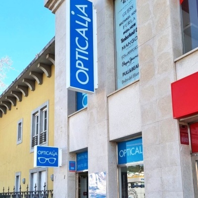 Loja Opticalia com placas azuis e brancas num edifício de pedra ao lado de um edifício amarelo