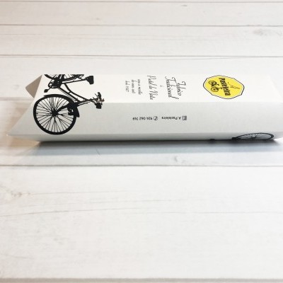 Embalagem branca com desenho de bicicleta preta e selo amarelo sobre superfície de madeira branca.