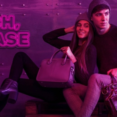 Homem e mulher com malas castanhas sentados contra parede cinzenta com luz rosa e texto neon 'BITCH, PLEASE'