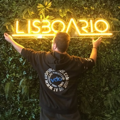 Letreiro de néon amarelo LISBOARIO numa parede verde com pessoa de costas vestindo camisola preta.