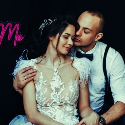 Casal em estúdio com fundo preto, mulher de vestido branco com renda e penas, homem com camisa branca e suspensórios, texto You & Me rosa e roxo