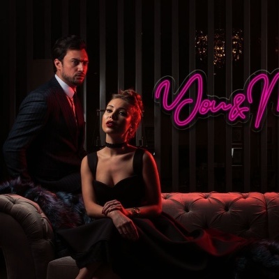 Homem e mulher vestidos formalmente sentados num sofá cinza com letreiro cor-de-rosa ao fundo