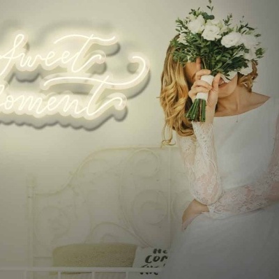 Noiva com vestido branco segurando ramo de flores diante de letreiro luminoso 'Sweet Moment?'