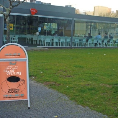 Placa publicitária laranja com texto sobre café num suporte metálico branco junto a relva e edifício de vidro ao fundo