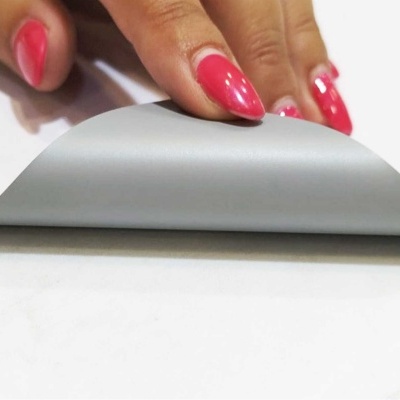 Mão com unhas cor de rosa a levantar uma folha flexível cinzenta e branca numa superfície branca.