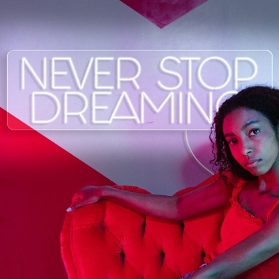 Jovem sentada num sofá vermelho com letreiro luminoso 'NEVER STOP DREAMING' ao fundo
