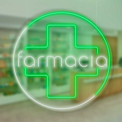 Letreiro de néon verde com cruz e texto 'farmácia' em interior de farmácia com prateleiras desfocadas