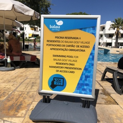 Placa informativa azul e amarela com regras da piscina do Balaia Golf Village junto a piscina e guarda-sóis brancos