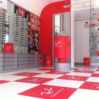 Loja com várias armações de óculos Ray-Ban exibidas em paredes de vidro e chão xadrez vermelho e branco com logotipo Ray-Ban