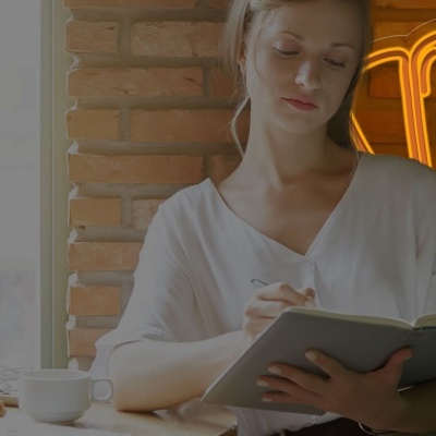 Mulher a escrever num caderno grande sentada num café com parede de tijolos e letreiro néon de croissant