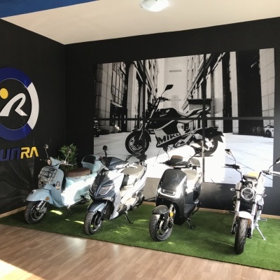 Quatro scooters elétricas em exposição interior com mural preto e branco e logo SUNRA na parede