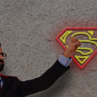 Homem em fato azul escuro e capa vermelha junto a emblema do super-homem iluminado na parede cinzenta