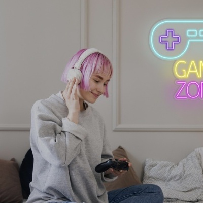Mulher jovem com cabelo rosa usando auscultadores e segurando comando de jogos num sofá com sinal de néon GAME ZONE na parede