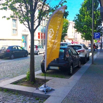 Bandeira publicitária amarela em tecido com base na calçada junto a árvore e carros estacionados