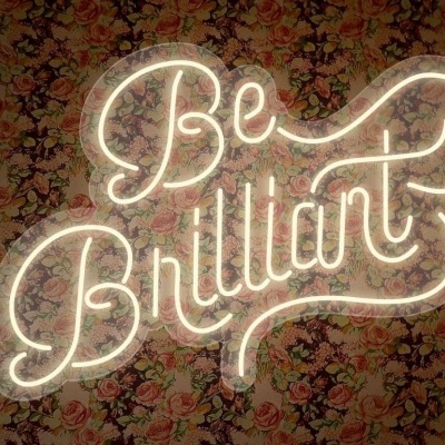 Letreiro de néon branco a dizer 'Be Brilliant' com fundo floral