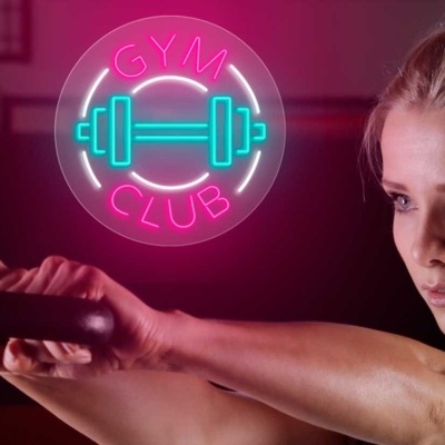 Mulher com kettlebell preto no ginásio com letreiro luminoso 'GYM CLUB'