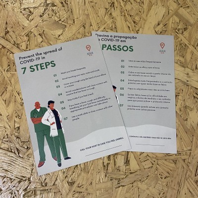 Folhetos informativos sobre prevenção da COVID-19 com texto em inglês e português, ilustração de profissionais de saúde, sobre mesa de madeira clara.