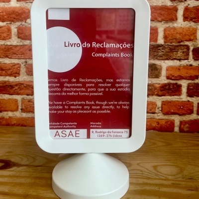 Expositor branco com informativo sobre Livro de Reclamações em mesa de madeira contra parede de tijolos