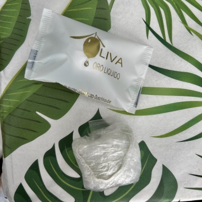 Embalagem branca OLIVA ORO LIQUIDO e pequena embalagem plástica transparente sobre tecido com padrão de folhas verdes