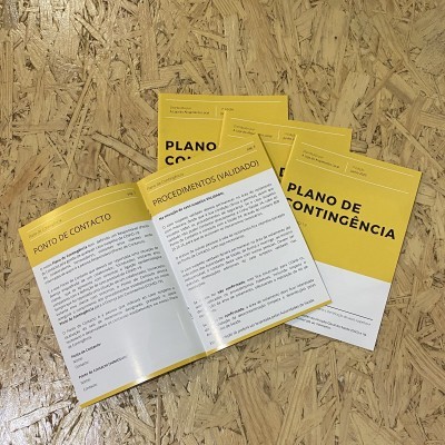 Livretos amarelos e brancos com texto em português 'PLANO DE CONTINGÊNCIA' sobre mesa de madeira