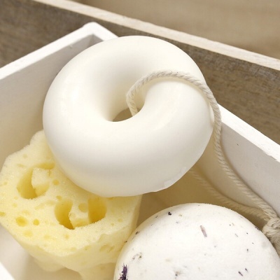 Produtos para banho: esponja amarela, sabão anel branco com cordão e bola branca com manchas dentro de caixa de madeira