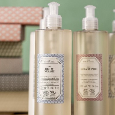 Três garrafas de plástico com rótulos coloridos e tampas de bomba, diante de caixas padronizadas.