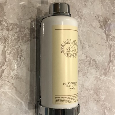 Frasco branco de loção corporal com tampa de bomba instalado em suporte metálico numa parede de mármore