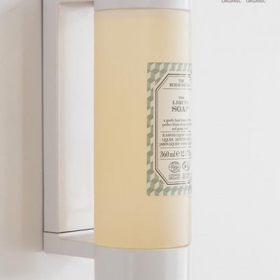 Dispenser de sabão líquido amarelo com tampa branca e etiqueta cinza, fixado na parede.