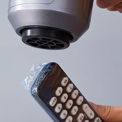 Telecomando preto com botões brancos coberto por plástico transparente sendo aquecido com secador de cabelo.
