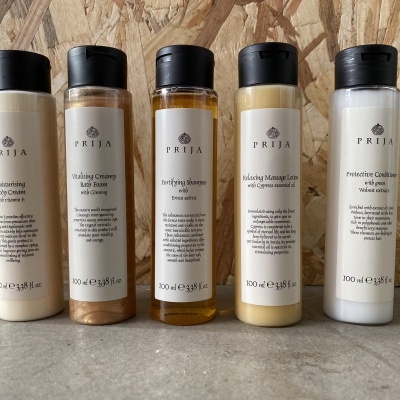 Cinco frascos de produtos PREIJA para cuidado corporal e capilar, com tampas pretas, cores variadas e rótulos claros, sobre superfície cinza e fundo de madeira.