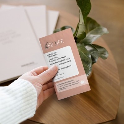 Pacote rosa de lenço higienizante para mãos SEE LIFE segurado à frente de planta e mesa de madeira