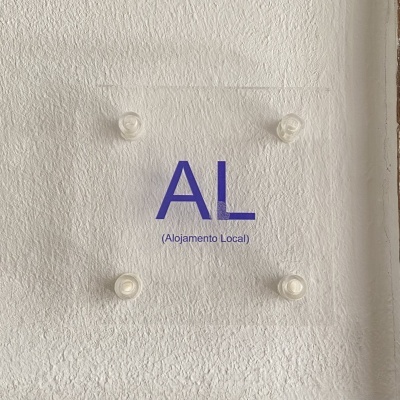 Placa transparente com o texto AL (Alojamento Local) em azul numa parede branca texturada