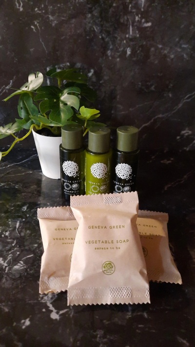 Frascos pequenos de shampoo e condicionador com embalagem verde e pacotes de sabonete vegeta em embalagem rosa clara, plantinha em vaso branco, sobre superfície escura mármore.