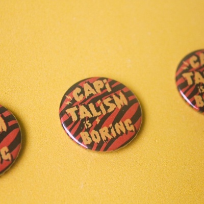 Três bottons com texto Capitalism is Boring sobre fundo amarelo.