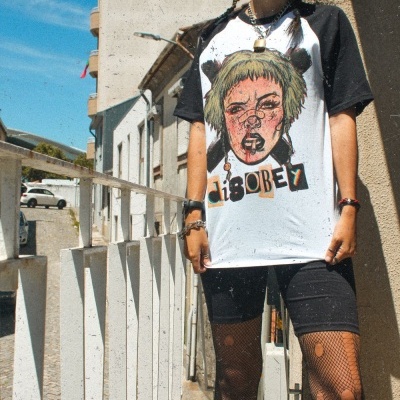 Pessoa com túnica branca e preta com ilustração, meias de rede rasgadas, boné e óculos de sol numa rua urbana