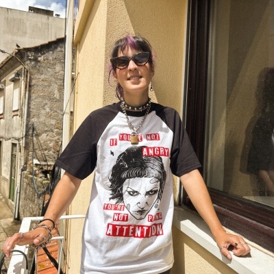 Homem de t-shirt preta e branca com estampa e texto, óculos escuros, pulseiras e colar de corrente a sorrir numa varanda com parede amarela.
