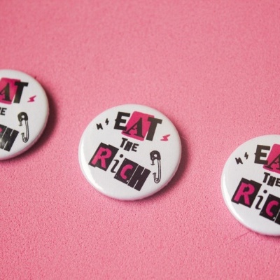 Três bottons redondos com texto EAT THE RICH em fundo rosa