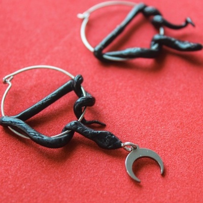 Brincos metálicos com detalhes pretos e pendente em forma de lua em fundo vermelho