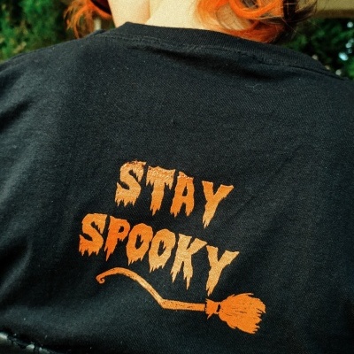 T-shirt preta com texto laranja STAY SPOOKY e desenho de vassoura