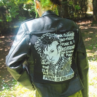 Casaco de couro preto com estampado punk e saia xadrez verde e amarelo vista das costas ao ar livre